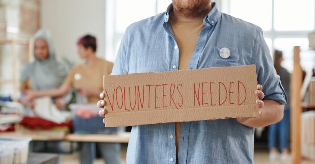 A man holds a 'Volunteers Needed' sign in a charitable organization setting, highlighting the need for help.